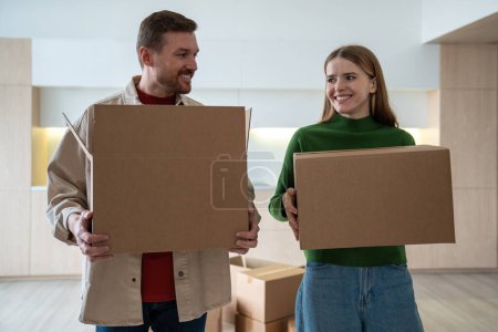 Smiling pleased spouses moving in new apartment house carrying cardboard boxes together. Happy family couple man and woman enjoying life changes. Relocation, mortgage, buying real estate, good mood.