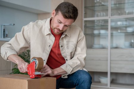 Concentrated man holding tape dispenser, sealing big cardboard box with adhesive scotch, packing belongings, preparing for moving day, relocation, transportation of belongings by shipping service