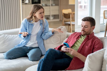 Indifferent man scrolls feed on social networks without paying attention to dissatisfied wife accusing of addiction of smartphone. Offended irritated woman asks husband ignore looking on screen phone