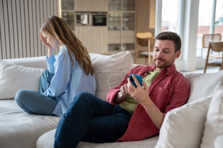 Frau weint, als der Mann zu Hause auf Couch sitzt. Mann ignoriert Gefühle seiner Frau beim Blick auf Smartphone-Bildschirm Ehepaare Missverständnisse in Beziehungen, eifersüchtiges Misstrauen. 