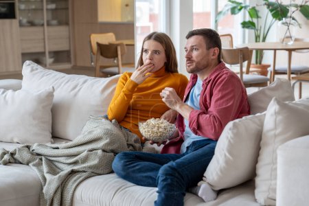 Amazed couple sitting on couch with popcorn, watching TV program, favourite show, film attentively, shocked, surprised by unexpected episode. Wife and husband spending weekends, pastime at home