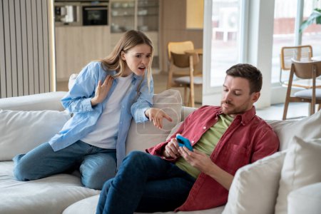 Indifferent ignoring man sits on sofa with mobile phone, playing games, scrolling social networks, reading news, keeping silence while offended irritated woman raises voice and shouts reproachfully