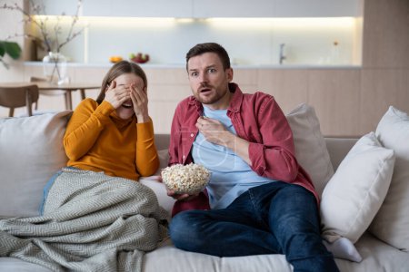 Couple spending time at home, watching TV, eating popcorn. Amazed intrigued husband looking with shoked interested glance. Embarrasing blushing wife closing eyes with hands of unexpected spicy episode
