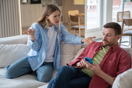 Indignant, offended, hysterical woman screams at husband sitting on sofa with cellphone. Couple family quarrel conflict misunderstanding discord. Indifferent man ignores dissatisfied wifes questions.