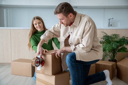 Middle-aged man holding tape dispenser, sealing big cardboard box with adhesive scotch, packing belongings, preparing for moving day, relocation. Cheerful pleasantly smiling woman helping, holding box