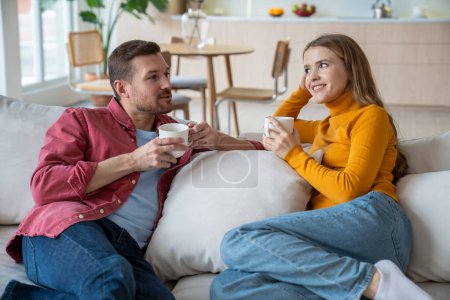 Foto de Pleased couple man woman talking sitting on home couch drinking coffee enjoying communication. Smiling wife flirting with husband listening him with interest. Happy marriage, harmony in relationship. - Imagen libre de derechos