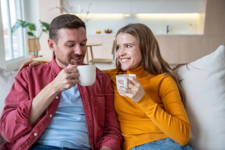Photo for Happy couple man and woman drinking tea coffee sitting on couch at home, enjoying talking tender hugging. Loving spouses wife husband bonding having nice conversation. Harmonious marriage relationship - Royalty Free Image