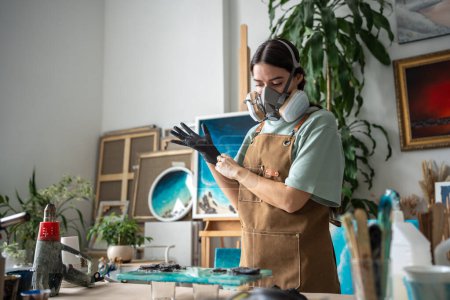 Creative female artist wearing respirator, latex gloves works with epoxy resin, crafting pieces on canvas in art studio. Manufacturing process of products made of liquid decorating material 