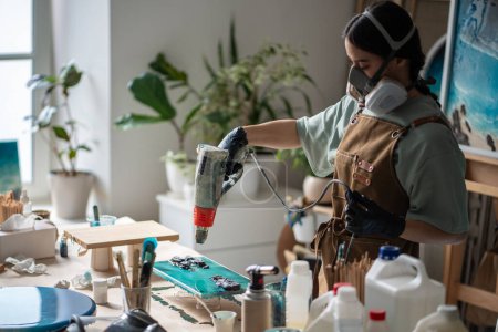 Artist girl works with flowable epoxy resin creates paintings in studio using construction thermal dryer. Woman creates handmade wooden serving boards in wood and epoxy resin with sea wave pattern.