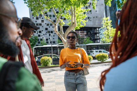 Interessante afrikanisch-amerikanische junge Frau mit Sonnenbrille, die auf der Straße steht und mit Freunden plaudert, Geschichten erzählt und tratscht. Gruppe schwarzer Studenten spricht nach Vorlesung im College im Freien
