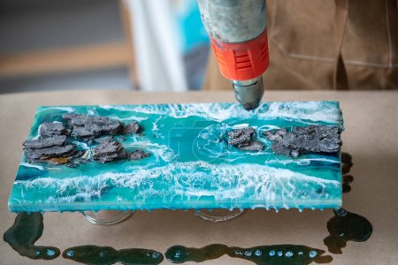 Artist working with liquid epoxy resin, creating sea, waves and rocks on two-part resin board, blowing a wooden painting with construction thermal dryer closeup. Masterclass in resin art, artwork, DIY