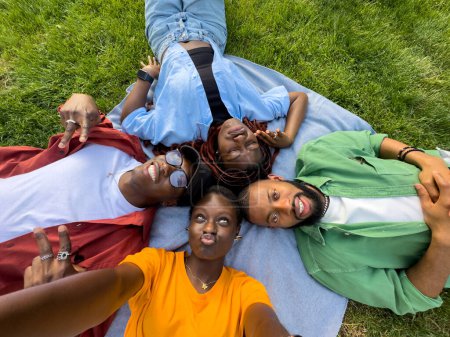 Happy group of african american friends girls and guys fool around, resting on grass, make selfie while grimacing. Satisfied four black students lying in lawn take picture on phone, have fun together.