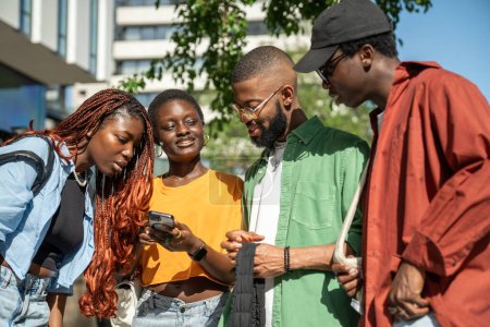 Interested stylish group of african young people students looks at smartphone while walking in street. Happy black girl shows social media post to friends on phone, bragging about high number of likes