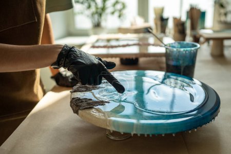 Resin art. Female artist applying epoxy resin with gloved fingers in art studio workshop. Closeup of woman wearing protective gloves removes excess uncured resin from painting with fingers. Fluid art.