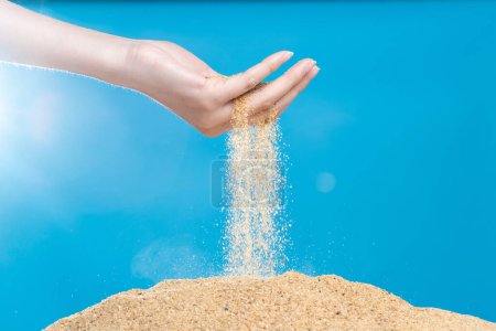 Photo for Hand releasing dropping sand. Fine Sand flowing pouring through fingers against blue background. Summer beach holiday vacation and time passing concept. Isolated high speed shutter - Royalty Free Image