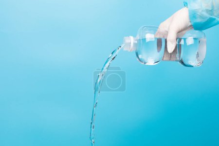 Téléchargez les photos : Saline medical cleaning normal solution plastic bottle in hand of nurse with glove, pouring water of normal saline in plastic bottle to clean sterile medical equipment over blue background isolated - en image libre de droit