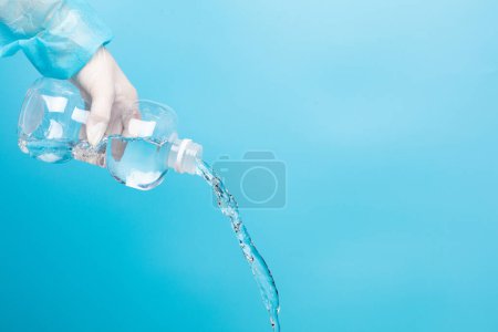Téléchargez les photos : Saline medical cleaning normal solution plastic bottle in hand of nurse with glove, pouring water of normal saline in plastic bottle to clean sterile medical equipment over blue background isolated - en image libre de droit