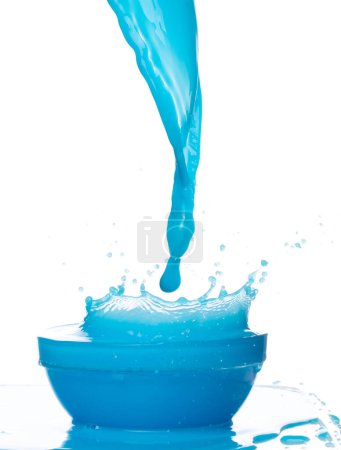 Téléchargez les photos : Versez de l'eau de peinture bleue dans une tasse de bol. Lotion liquide bleue hydratant cosmétique verser flotteur dans l'air milieu. L'explosion d'une boisson cocktail bleue tremble. Fond blanc isolé obturateur à grande vitesse - en image libre de droit