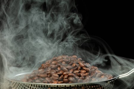 Téléchargez les photos : Graines de café torréfiées avec fumée. Haricots Café torréfié sur pile et fumée chaude lever en forme de ligne. Grain de café torréfié créer un arôme parfumé. Fond noir isolé - en image libre de droit