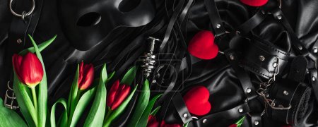 Explora el mundo del juego pervertido. Vista superior de látigos kit de cuero bdsm, esposas, máscara y cadena contra la seda negro. tulipanes
