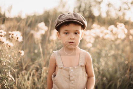 Téléchargez les photos : A boy is playing on a farm or ranch field, resting. Portrait of a child boy in a cap and overalls. Childhood - en image libre de droit