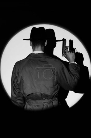 Foto de Una silueta oscura de un detective con un abrigo y un sombrero con una pistola en las manos al estilo negro. Un retrato dramático en el estilo de las películas de detectives de la década de 1950. La silueta de un espía en un círculo de luz. monocromo - Imagen libre de derechos