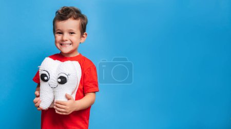A smiling boy with healthy teeth holds a plush tooth in his hands on a blue isolated background. Oral hygiene. Pediatric dentistry. Prevention of caries. A place for your text
