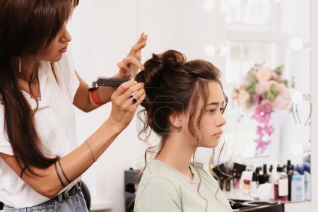Photo for Shooting in a beauty salon. The hair master corrects the hairstyle of a young dark-haired girl with the help of a hair brush - Royalty Free Image