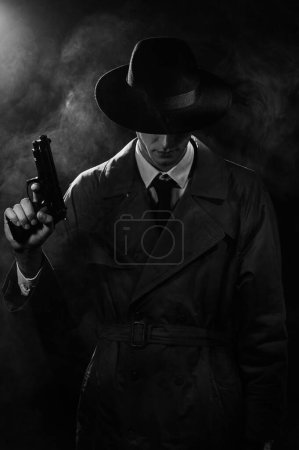 Foto de Una silueta oscura de un detective con un abrigo y un sombrero con una pistola en las manos al estilo Noir. La cabeza está baja. Un dramático retrato negro al estilo de los detectives de los años 60. monocromo - Imagen libre de derechos