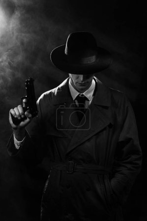 Foto de Una silueta oscura de un detective con un abrigo y un sombrero con una pistola en las manos al estilo Noir. La cabeza está baja. Un dramático retrato negro al estilo de los detectives de los años 60 - Imagen libre de derechos