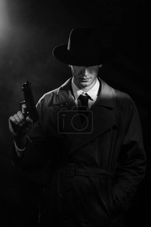 Foto de Una silueta oscura de un detective con un abrigo y un sombrero con una pistola en las manos al estilo Noir. La cabeza está baja. Un dramático retrato negro al estilo de los detectives de los años 50 - Imagen libre de derechos