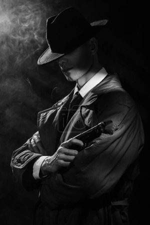 Foto de Una silueta oscura de un hombre con un impermeable y un sombrero con una pistola en las manos al estilo del negro del crimen. Un dramático retrato negro al estilo de los detectives de los años 50 - Imagen libre de derechos