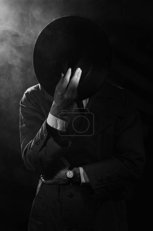 Foto de Una silueta oscura de un hombre con un impermeable y un sombrero al estilo del negro del crimen. Retrato dramático de negro al estilo de los detectives de los años 50. Hay humo en la foto. - Imagen libre de derechos