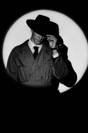 Foto de A dark silhouette of a male detective in a coat and hat in the noir style. A dramatic portrait in the style of detective films of the 1950s. A silhouette in a circle of light, like Agent 007 - Imagen libre de derechos
