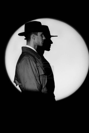 Foto de A dark silhouette of a male detective in a coat and hat in the noir style. A dramatic portrait in the style of detective films of the 1950s. The silhouette of a spy in a circle of light, like Agent - Imagen libre de derechos