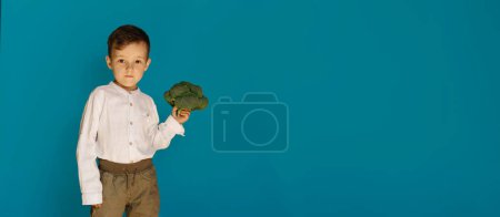 A studio shot of a smiling boy holding fresh broccoli on a blue background with a copy of the space. The concept of healthy baby food