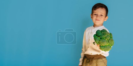 A studio shot of a smiling boy holding fresh broccoli on a blue background with a copy of the space. The concept of healthy baby food