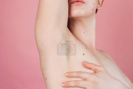 A young beautiful shirtless woman showing off her hairy armpits on an isolated pink background