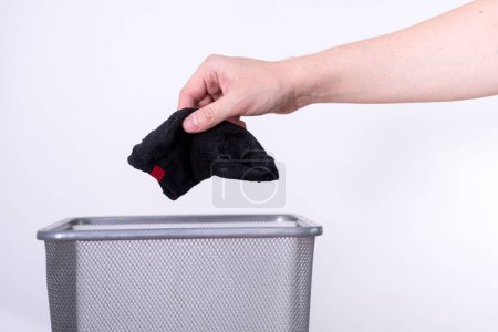 Foto de Un hombre está tirando un calcetín viejo a un cubo de basura con la mano contra un fondo blanco. El concepto de ropa vieja. - Imagen libre de derechos