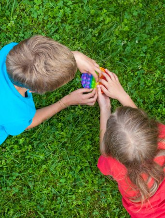 Kinder, die mit rundem Regenbogen spielen, lassen es (Popit-) Spielzeug auf dem Rasen knallen. Junge und Mädchen. Neues Fidget Trendspielzeug. Antistress-Spielzeug. Einfache Grübchen.