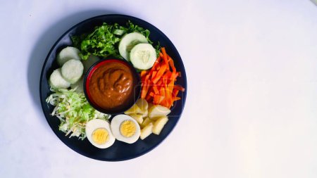 Gado-gado servido en un plato, aislado blanco, ensalada indonesia que contiene verduras hervidas y patatas, huevos cocidos, tempeh tofu frito, lontong y chips de melinjo, servido con salsa de cacahuete y anacardo. 