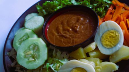 Gado-gado servido en un plato, aislado blanco, ensalada indonesia que contiene verduras hervidas y patatas, huevos cocidos, tempeh tofu frito, lontong y chips de melinjo, servido con salsa de cacahuete y anacardo. 