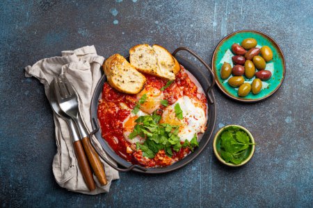 Photo for Middle Eastern and Maghrebi healthy dish Shakshouka made of eggs and tomato sauce served in pan with toasts, fresh cilantro and olives on rustic concrete background table from above - Royalty Free Image