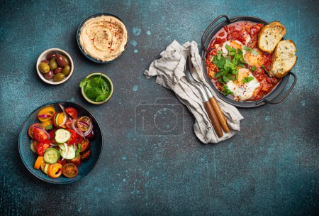 Photo for Middle Eastern traditional breakfast or brunch with eggs Shakshouka in pan with toasts, fresh vegetables salad, hummus and olives on rustic concrete background table from above, space for text - Royalty Free Image