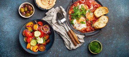 Photo for Middle Eastern traditional breakfast or brunch with eggs Shakshouka in pan with toasts, fresh vegetables salad, hummus and olives on rustic concrete background table from above - Royalty Free Image