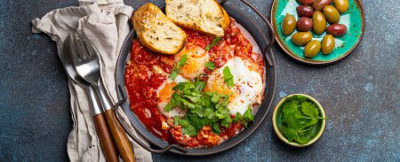 Photo for Middle Eastern and Maghrebi healthy dish Shakshouka made of eggs and tomato sauce served in pan with toasts, fresh cilantro and olives on rustic concrete background table from above - Royalty Free Image