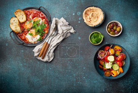 Photo for Middle Eastern traditional breakfast or brunch with eggs Shakshouka in pan with toasts, fresh vegetables salad, hummus and olives on rustic concrete background table from above, space for text - Royalty Free Image