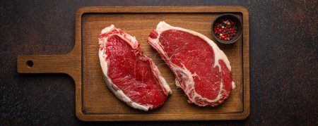 Two raw uncooked meat beef rib eye marbled steaks on wooden cutting board with seasonings on dark rustic background ready to be grilled from above, preparing dinner with meat