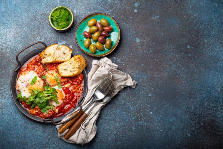 Photo for Middle Eastern and Maghrebi healthy dish Shakshouka made of eggs and tomato sauce served in pan with toasts, fresh cilantro and olives on rustic concrete background table from above, space for text - Royalty Free Image