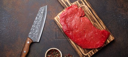 Roh rohes rundes Rindfleisch-Steak auf Holzschneidebrett mit großem Küchenmesser und Pfeffer auf dunkelbraunem rustikalem Stein Hintergrundansicht, Kochfleisch-Steakkonzept
